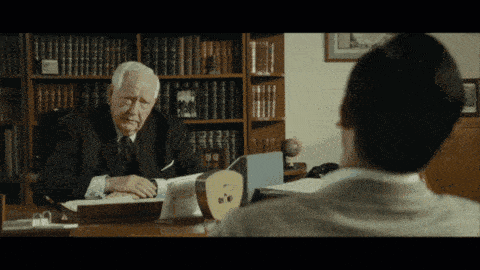 a man in a suit sits at a desk with a clock on it|833x468.5625